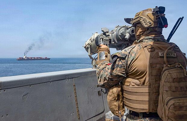Im Vordergrund ein Soldat auf einem Schiff, der einen Öltanker im Hintergrund mit einem Fernrohr ins Visier nimmt