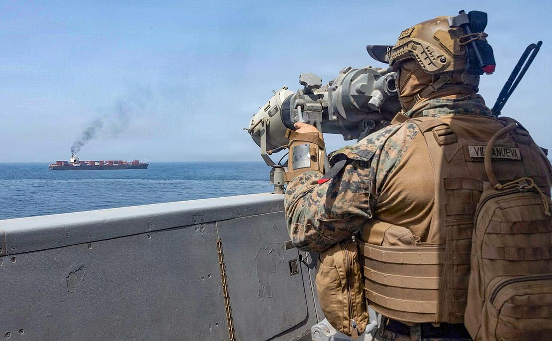 Im Vordergrund ein Soldat auf einem Schiff, der einen Öltanker im Hintergrund mit einem Fernrohr ins Visier nimmt