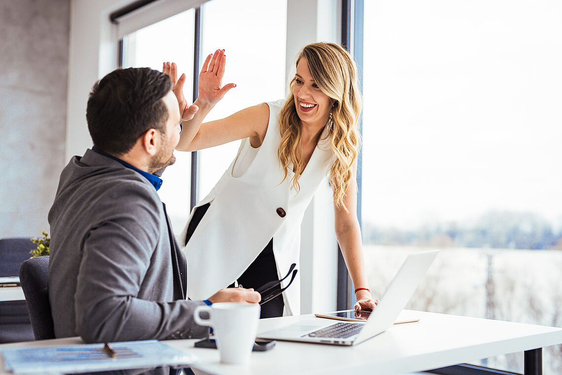 Ein Mann - sitzend am Schreibtisch - und eine Frau - stehend daneben - klatschen freudig ab