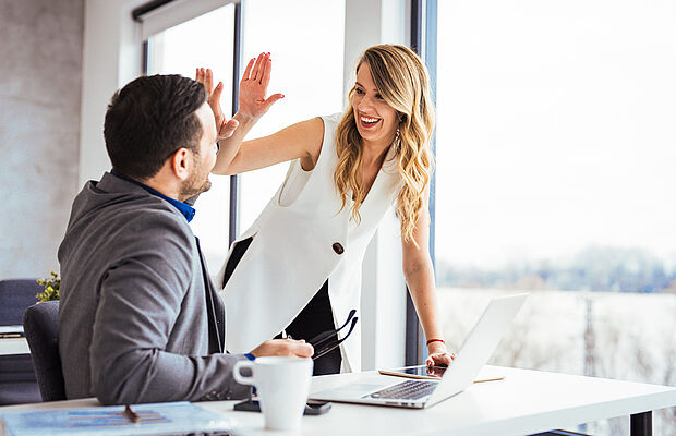 Ein Mann - sitzend am Schreibtisch - und eine Frau - stehend daneben - klatschen freudig ab