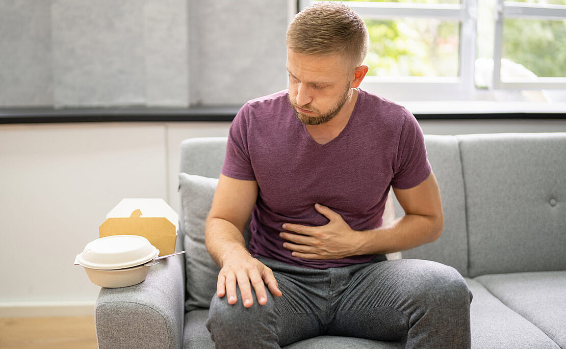 Mann auf Sofa mit einer Hand am Bauch, Völlegefühl, Blähungen