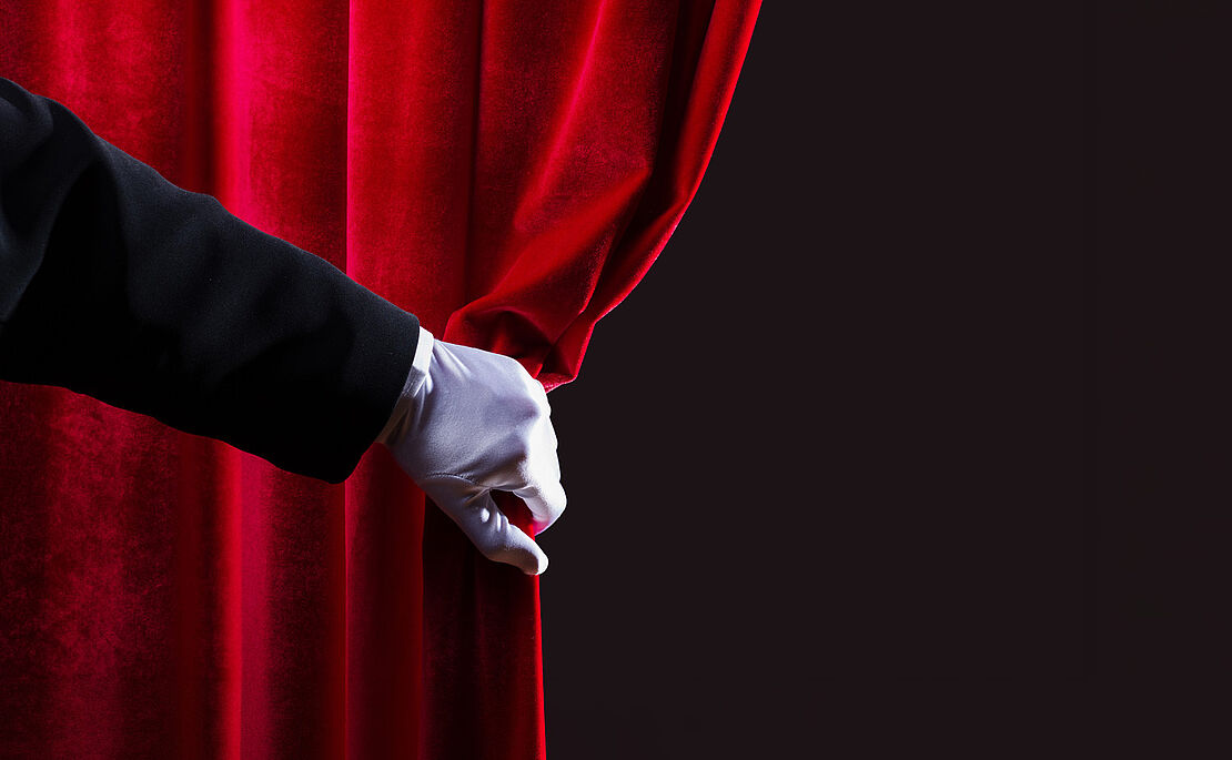 Ein roter Vorhang, der von einer Hand im weißen Handschuh beiseite gezogen wird, dahinter schwarzer Raum