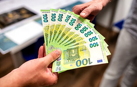 Eine Hand mit einem Fächer von 100 Euro Scheinen, zweite Hand greift zu den Scheinen