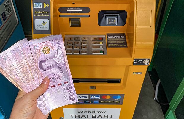 Eine Frauenhand mit vielen thailandischen Geldscheinen in der Hand vor einem gelben Bankomat