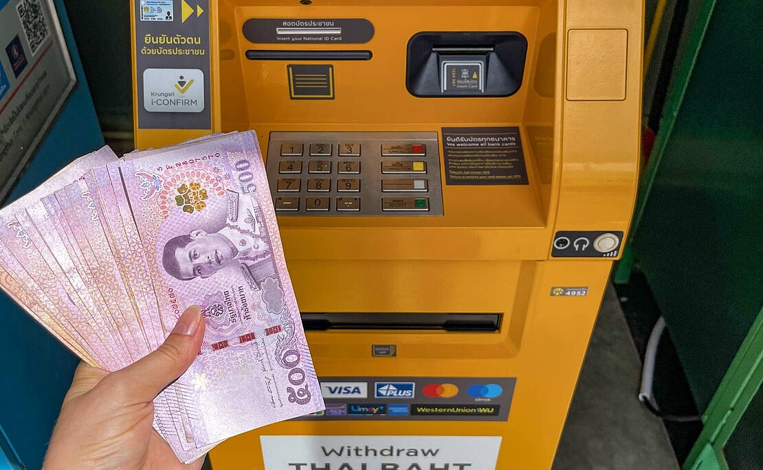Eine Frauenhand mit vielen thailandischen Geldscheinen in der Hand vor einem gelben Bankomat