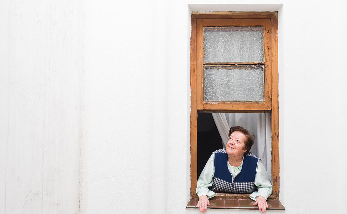 Ältere Frau, die aus dem Fenster Richtung Himmel sieht, die Hände auf das Fensterbrett gestützt