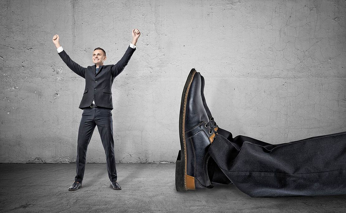 Mann, der vor Freude beide Arme in die Höhe streckt, rechts im Bild ein übergroßes Schienbein mit übergroßen Schuhen