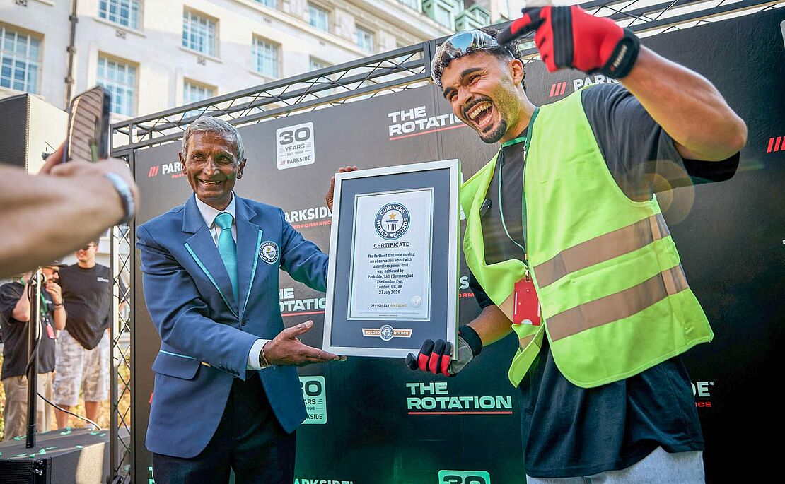 Zwei Männer, die zwischen sich ein gerahmtes Dokument halten, der rechte Mann in Bauarbeitermontur, der sich freut und auf das Bild zeigt
