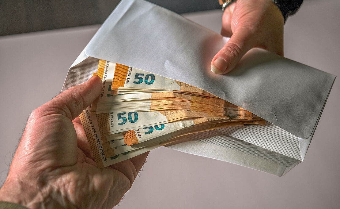 Zwei Hände bei Geldübergabe, viele 50-Euro-Scheine schauen aus einem Kuvert