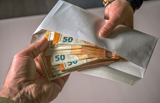 Zwei Hände bei Geldübergabe, viele 50-Euro-Scheine schauen aus einem Kuvert