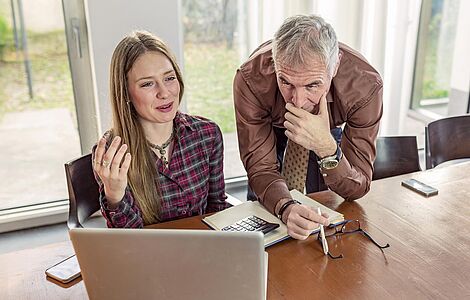 Junge Frau vor PC, älterer grauhaariger Mann daneben über den Tisch gebeugt