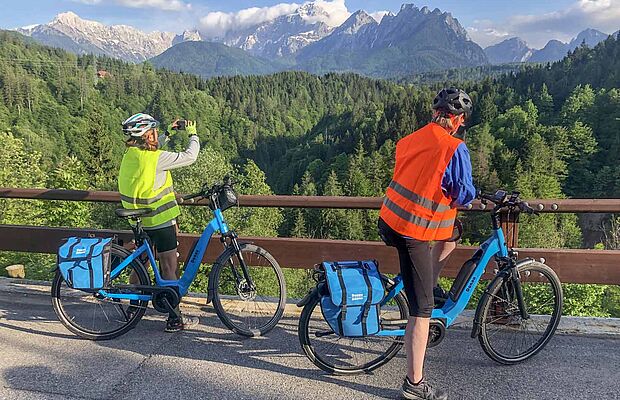 Mann und Frau auf E-Bike, Frau fotografiert die Landschaft bei Tarvis mit Blick auf die Julischen Alpen