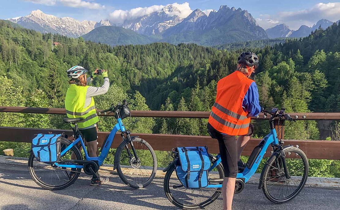 Mann und Frau auf E-Bike, Frau fotografiert die Landschaft bei Tarvis mit Blick auf die Julischen Alpen