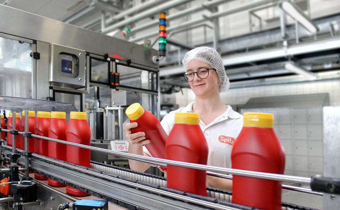 Junge Frau mit Haarnetz und weißem Shirt am Fließband mit Ketchupflaschen