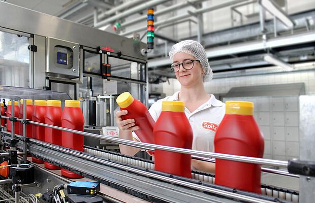 Junge Frau mit Haarnetz und weißem Shirt am Fließband mit Ketchupflaschen