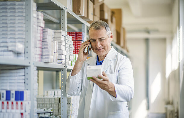 Onlineapotheken, Mitarbeiter telefoniert und hält eine Medikamentenpackung in der Hand, im Hintergrund ein hohes Regal mit Medikamenten