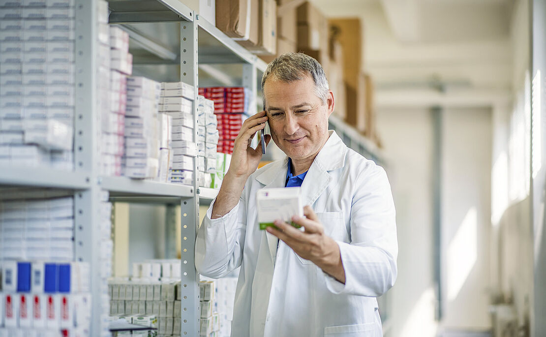 Onlineapotheken, Mitarbeiter telefoniert und hält eine Medikamentenpackung in der Hand, im Hintergrund ein hohes Regal mit Medikamenten
