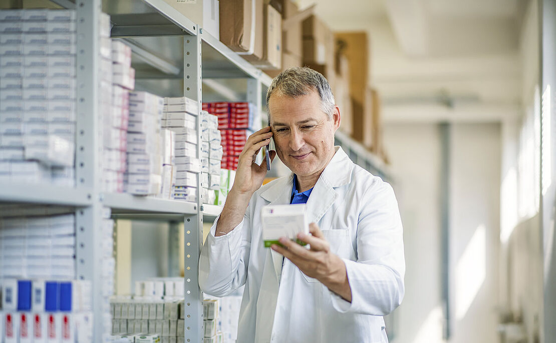 Onlineapotheken, Mitarbeiter telefoniert und hält eine Medikamentenpackung in der Hand, im Hintergrund ein hohes Regal mit Medikamenten