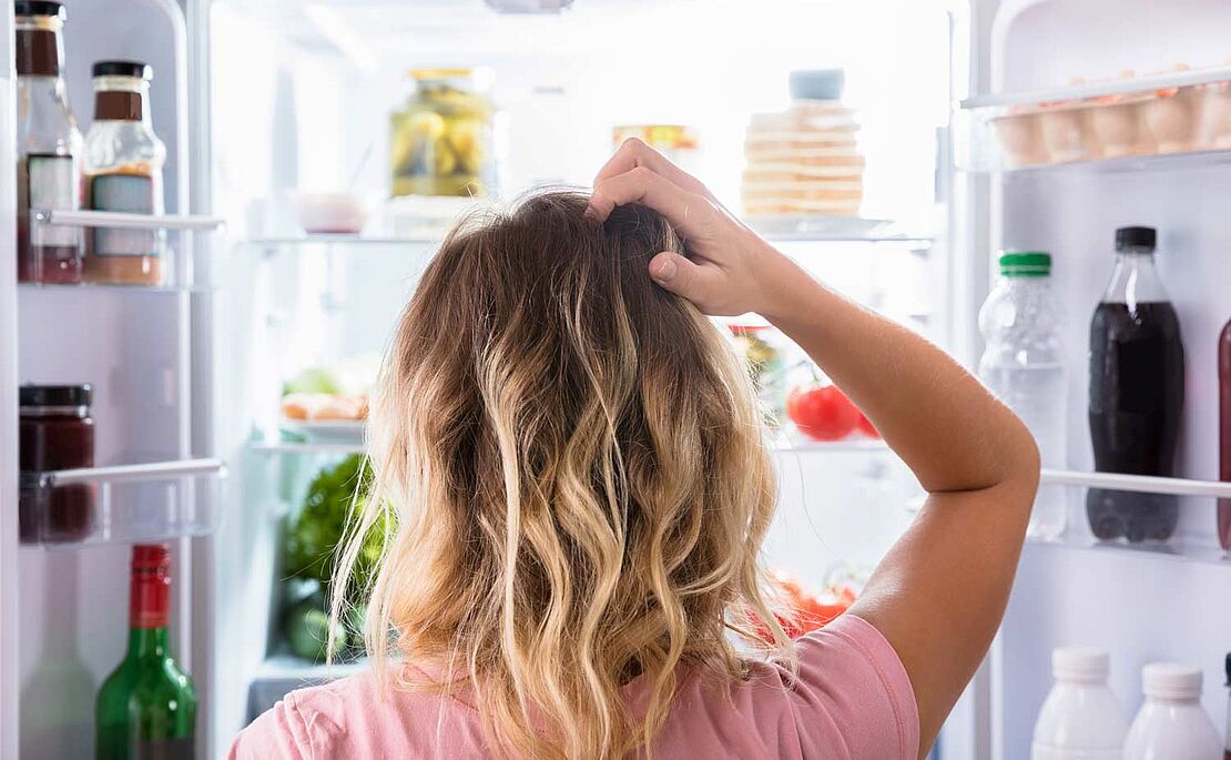 Frau, die sich ihre langen blonden Haare vor einem offenen Kühlschrank mit einer Vielzahl an Lebensmitteln rauft