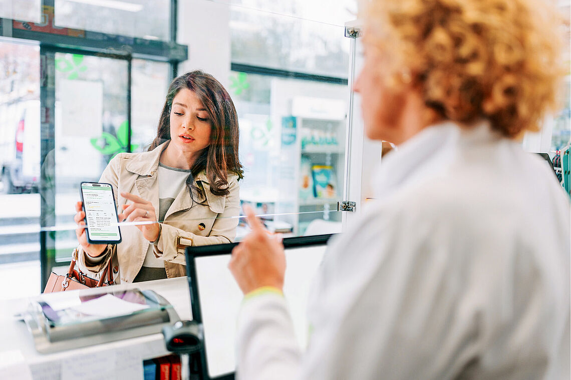 Junge Dame zeigt einer Apothekerin etwas auf ihrem Handy