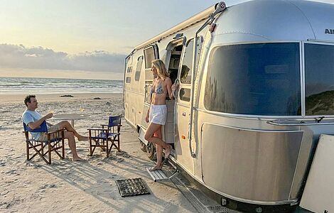 Silbernes Wohnmobil, geparkt am Strand, eine Frau im Bikini tritt gerade aus der Tür, ein Mann sitzt davor an Tisch mit zwei Sesseln