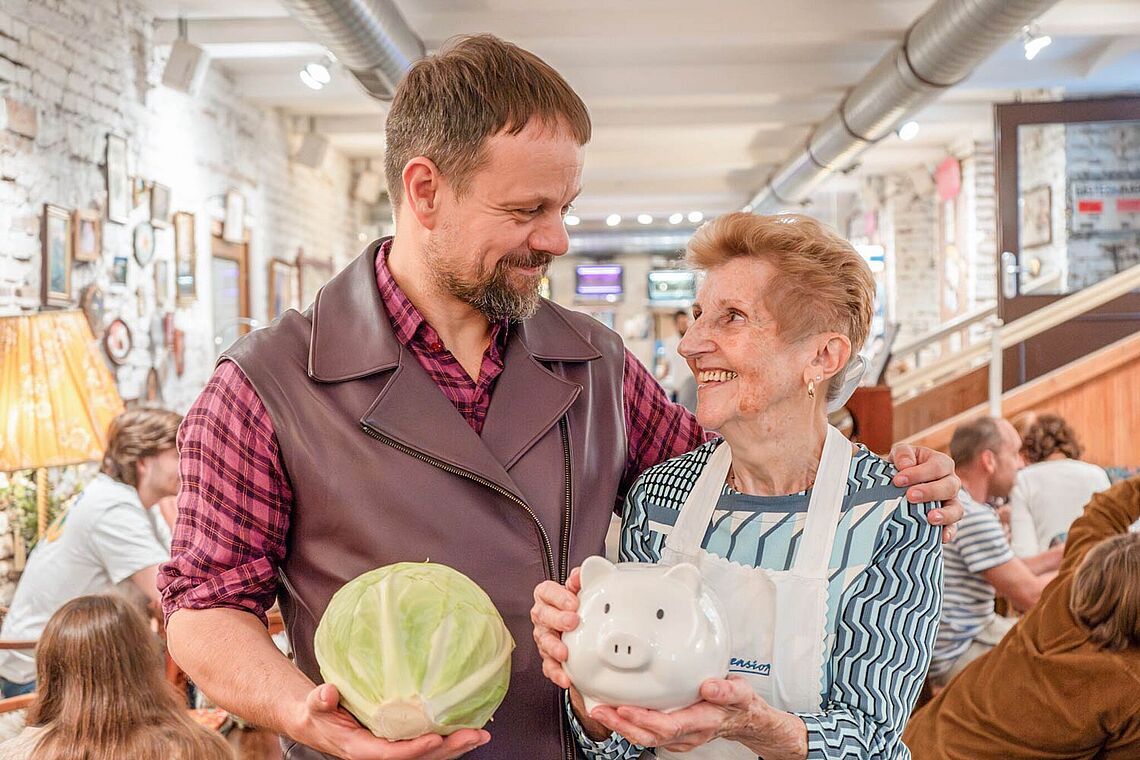 Mann mit Krautkopf in der Hand, ältere Dame mit Sparschwein in der Hand, beide sehen sich an