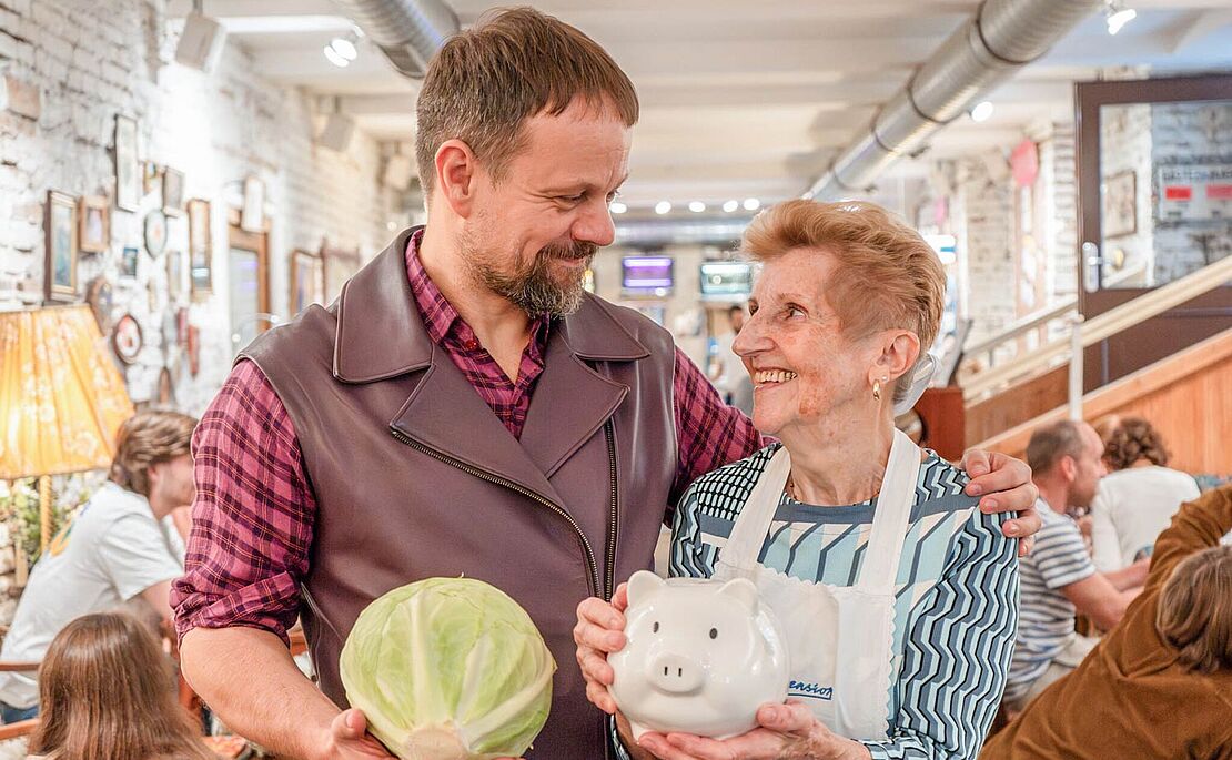 Mann mit Krautkopf in der Hand, ältere Dame mit Sparschwein in der Hand, beide sehen sich an