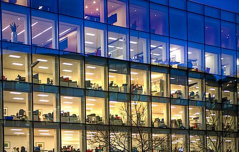 Bürogebäude mit vielen hell erleuchteten Fenstern und Mitarbeitern, die noch spät arbeiten