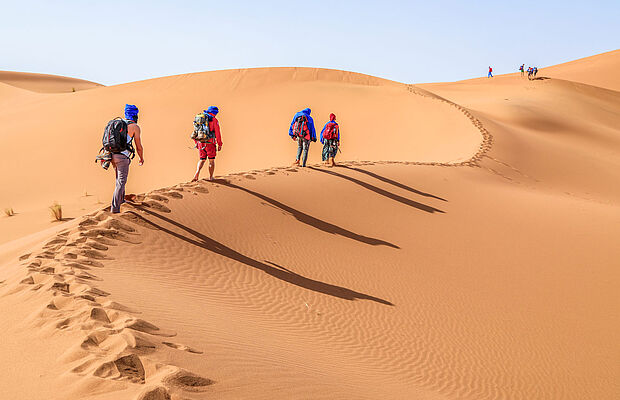 Vier Personen gehen mit Rucksack und Kleidung zum Schutz vor der Sonne durch die Wüste, Sanddünen im Erg Chegaga bei Zagora in Marokko