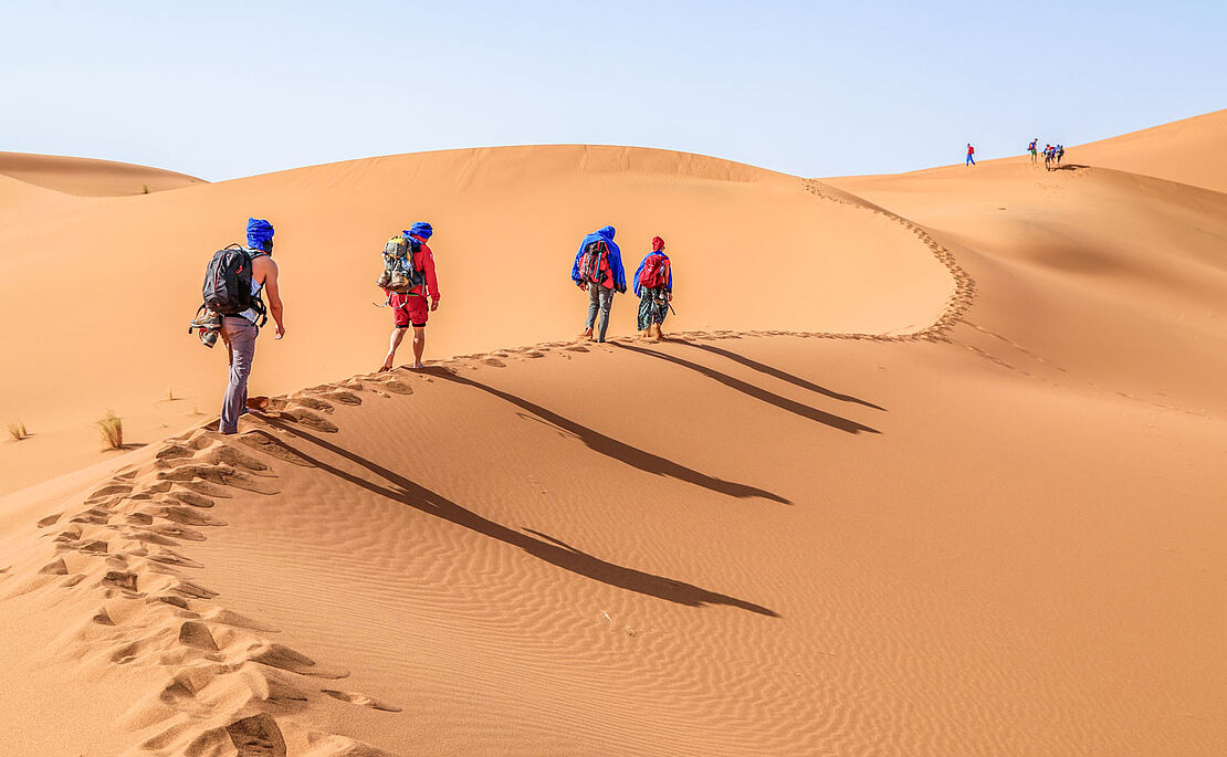 Vier Personen gehen mit Rucksack und Kleidung zum Schutz vor der Sonne durch die Wüste, Sanddünen im Erg Chegaga bei Zagora in Marokko