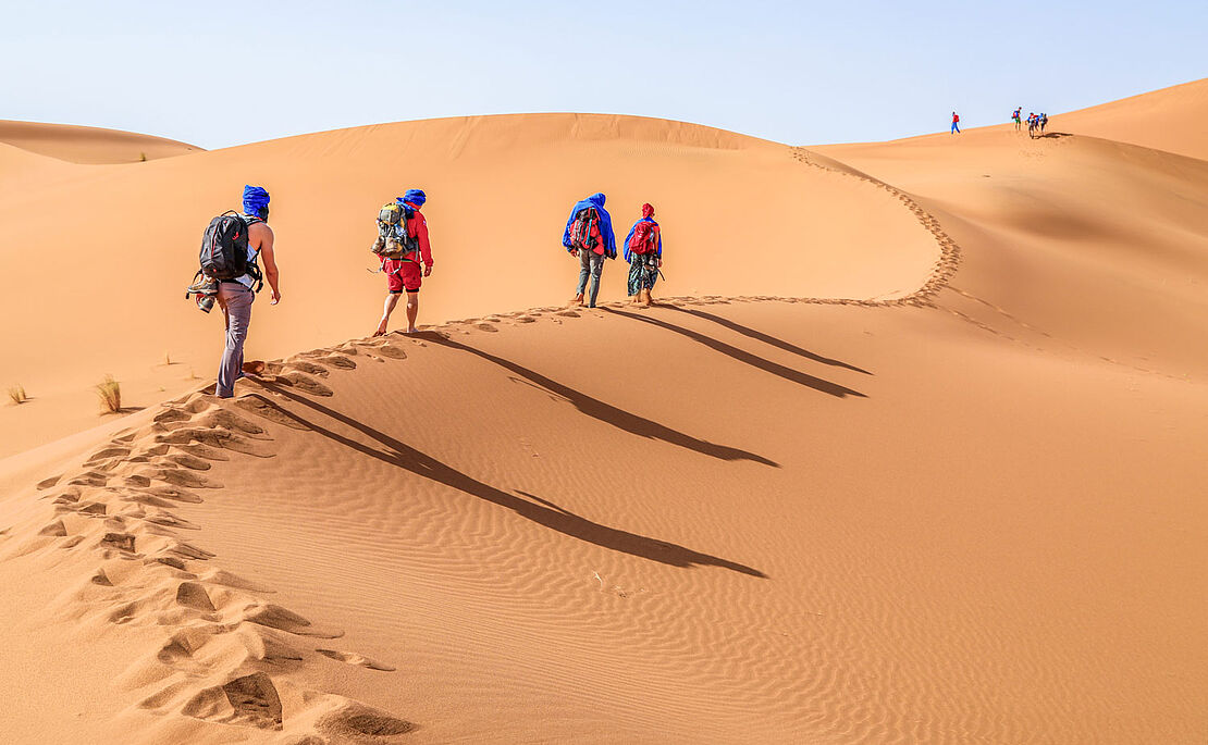 Vier Personen gehen mit Rucksack und Kleidung zum Schutz vor der Sonne durch die Wüste, Sanddünen im Erg Chegaga bei Zagora in Marokko