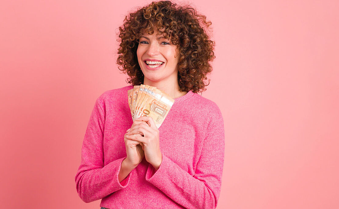 Aufgeregte junge Frau in pinkem Outfit mit lockigem kurzen Haar zeigt einige 50-Euro-Banknoten