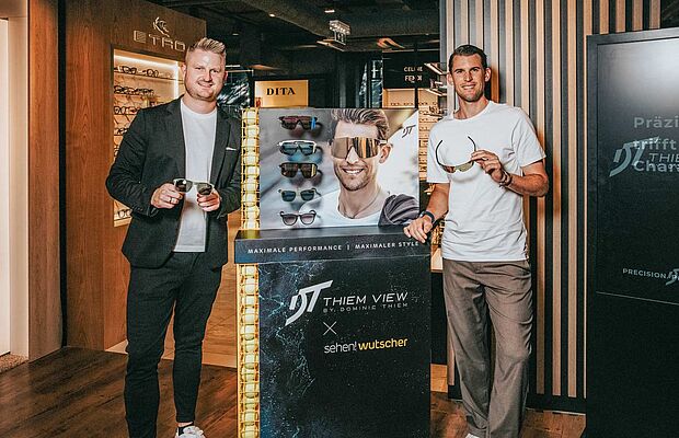 Fritz Wutscher jr. und Dominic Thiem vor einem Brillengeschäft, jeder hält eine Sonnenbrille in der Hand, in ihrer Mitte ein Werbeständer