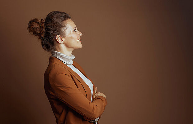 Frau mit Dutt hellem Rollkragenpulli und braunem Blazer seitwärts gerichtet, blickt nach oben