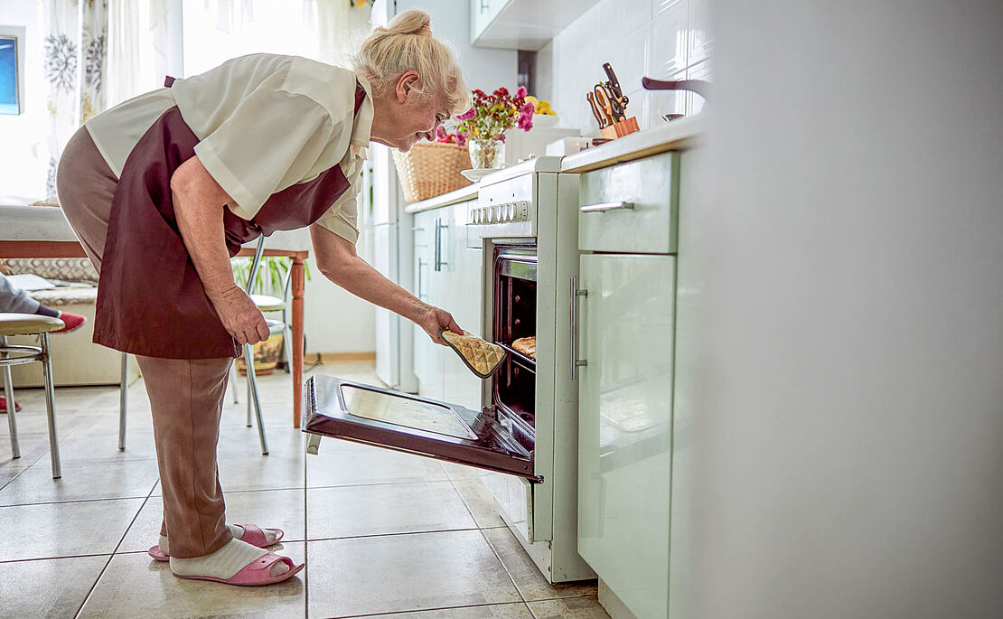 Frau im gehobenen Alter, die in einer Küche mit Kochschürze etwas aus dem Backrohr nimmt
