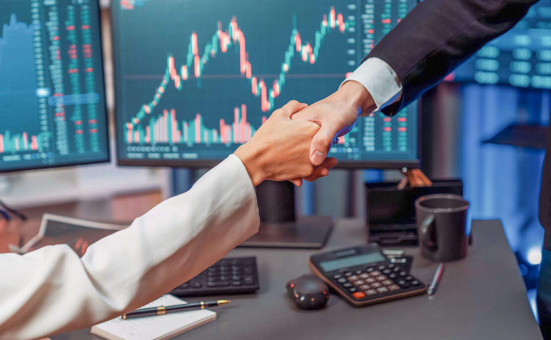 Handshake zwischen Frau und Mann, im Hintergrund Bildschirme mit Chartverläufen