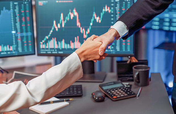 Handshake zwischen Frau und Mann, im Hintergrund Bildschirme mit Chartverläufen
