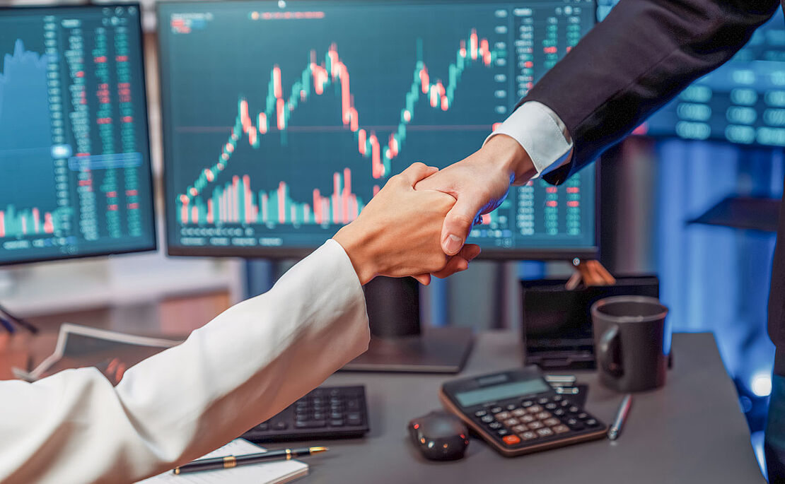 Handshake zwischen Frau und Mann, im Hintergrund Bildschirme mit Chartverläufen