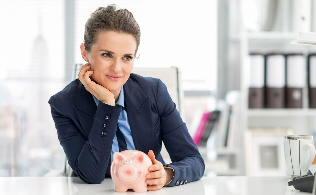 Businessfrau an einem Tisch sitzend, ein Sparschwein vor ihr haltend