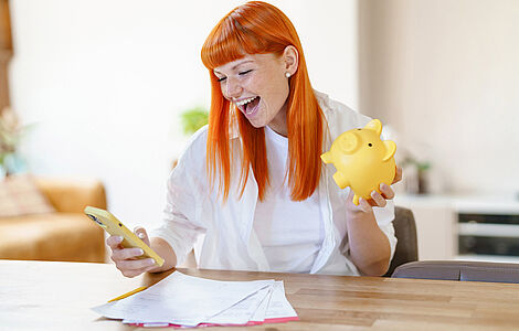 Lachende Frau mit roten Haaren und einem Handy und einem Sparschwein in der Hand