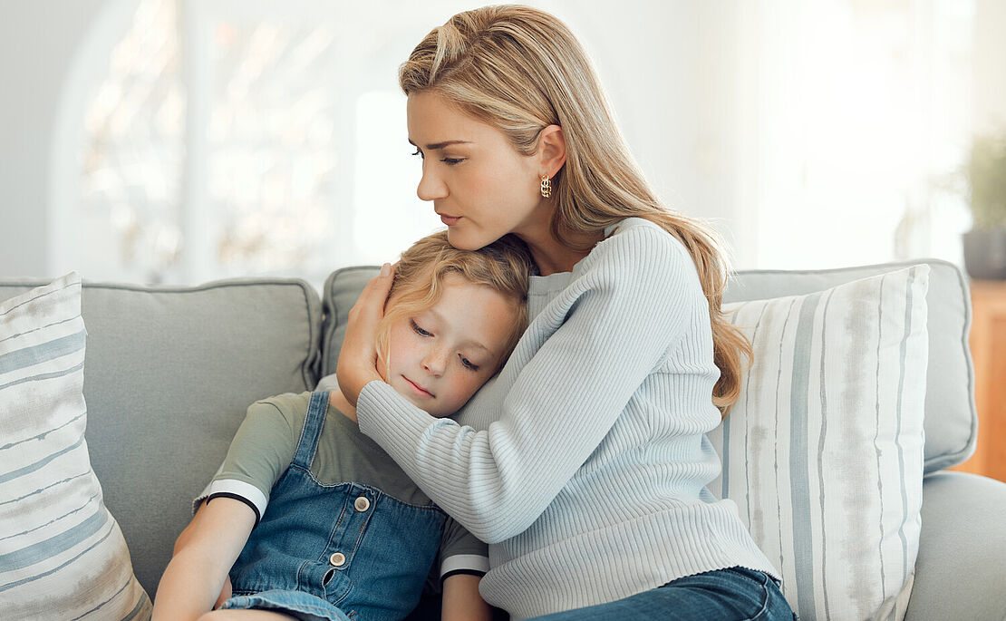 Mutter sitzt mit Tochter auf Couch und hält sie im Arm
