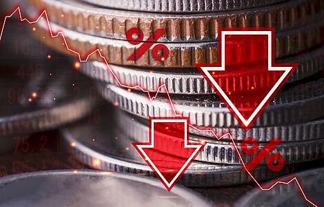 Herangezoomtes Foto eines Münzstapels, darüber sind mit Bildbearbeitung zwei rote nach unten zeigende Pfeile, zwei Prozentzeichen und eine fallende Kurskurve gelegt