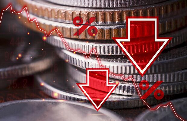 Herangezoomtes Foto eines Münzstapels, darüber sind mit Bildbearbeitung zwei rote nach unten zeigende Pfeile, zwei Prozentzeichen und eine fallende Kurskurve gelegt