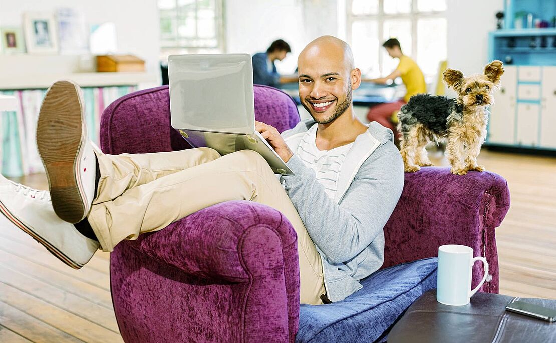 Ein Mann auf einem lila Fauteuil sitzend, die Beine über die Lehne geschlagen, ein Laptop auf dem Schoß, ein Hündchen auf der anderen Lehne stehend, im Hintergrund ein Wohnzimmer mit anderen Personen