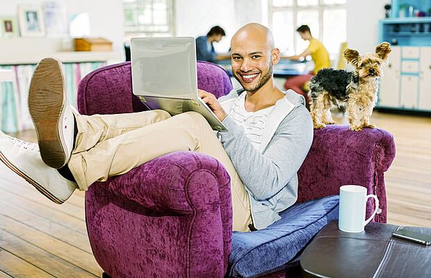 Ein Mann auf einem lila Fauteuil sitzend, die Beine über die Lehne geschlagen, ein Laptop auf dem Schoß, ein Hündchen auf der anderen Lehne stehend, im Hintergrund ein Wohnzimmer mit anderen Personen