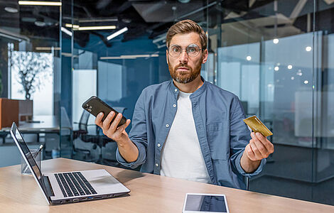 Mann mit Brille und Vollbart hält in einer Hand das Handy, in der anderen eine Kreditkarte und blickt verwundert