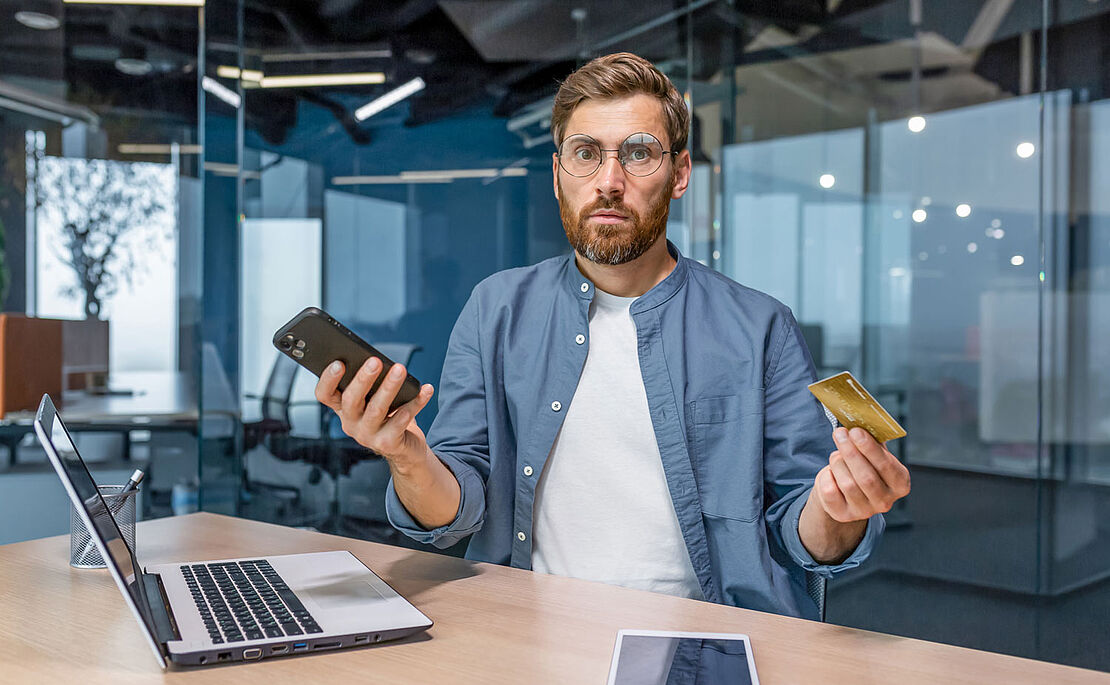 Mann mit Brille und Vollbart hält in einer Hand das Handy, in der anderen eine Kreditkarte und blickt verwundert