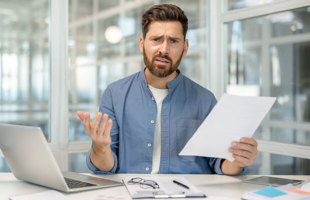 Mann vor PC und mit Papier in der Hand, der fragend und verständnislos in die Kamera schaut