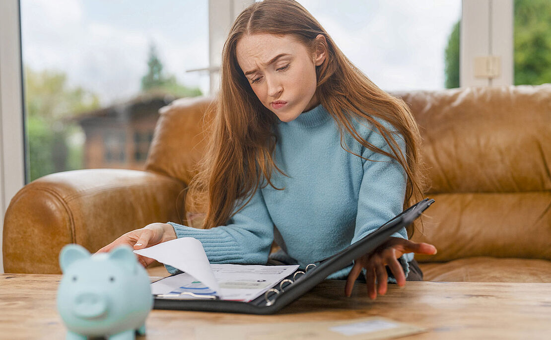 Junge Frau bei Durchsicht einer Mappe, vor ihr ein Sparschwein